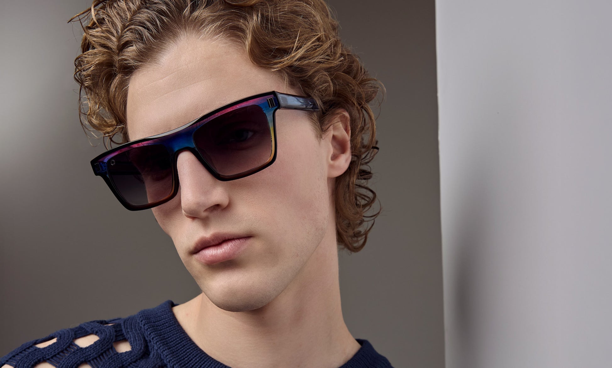 A close-up of a man with curly light brown hair wearing angular sunglasses with a dark blue, purple, and pink iridescent frame and dark lenses, paired with a navy knit sweater.