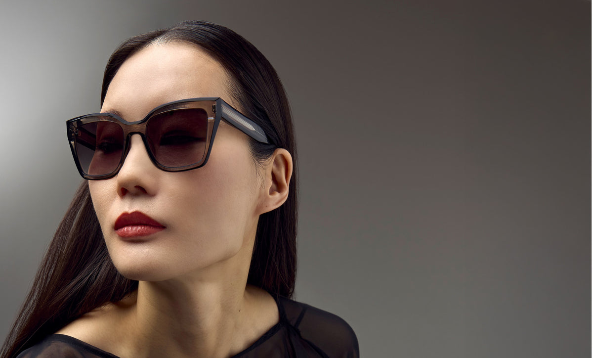 A woman with long dark hair wearing stylish cat-eye sunglasses with dark brown lenses and a black frame, set against a solid gray studio background.