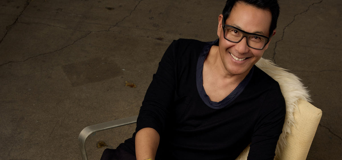 A smiling man with glasses wearing a black long-sleeve shirt with a dark blue collar, seated in a cream-colored armchair with a white faux-fur accent, against a dark concrete floor background.