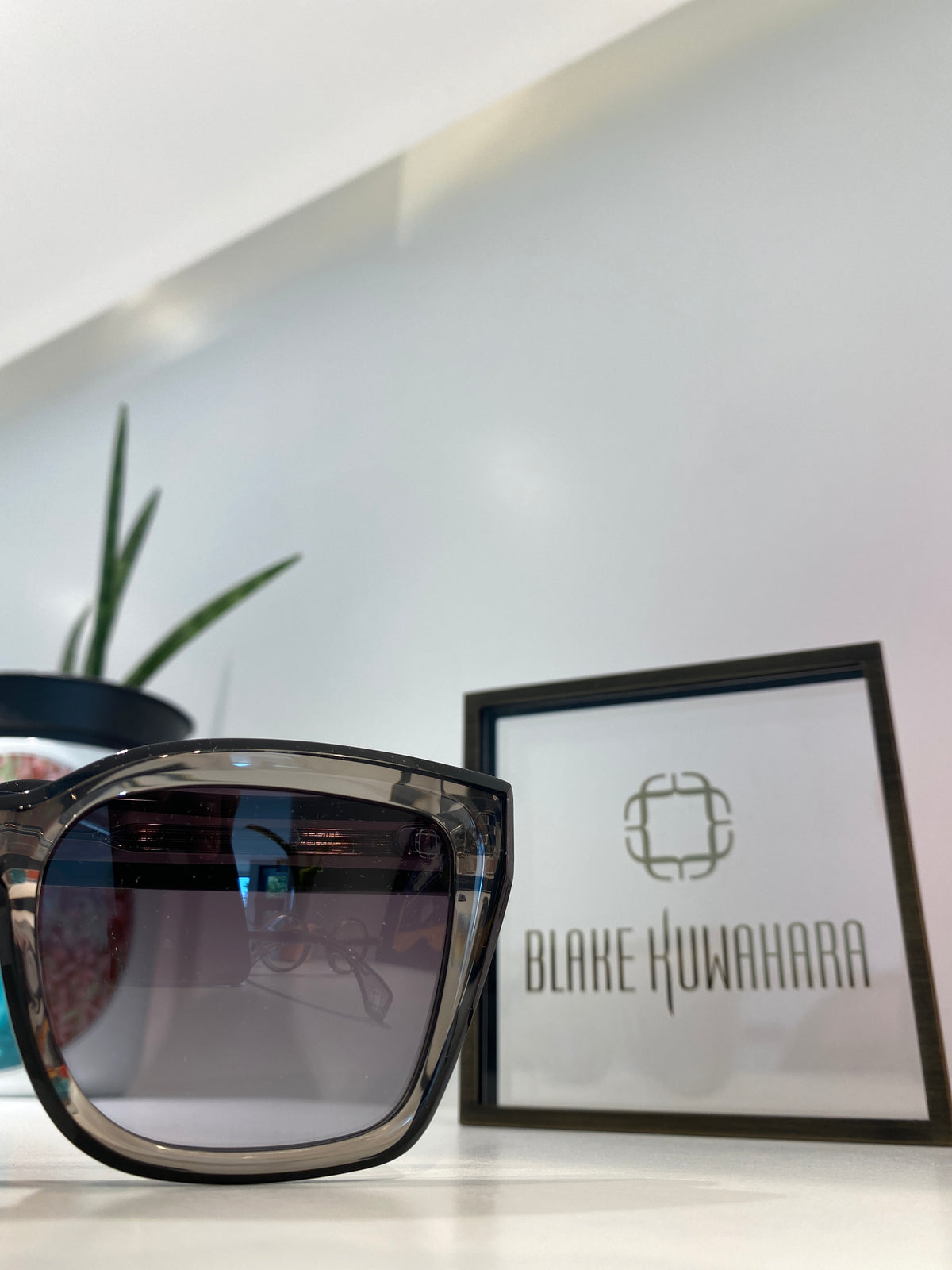 A pair of oversized square sunglasses with a translucent smoky gray frame and dark tinted lenses, displayed on a white surface next to a framed sign reading Blake Kuwahara.