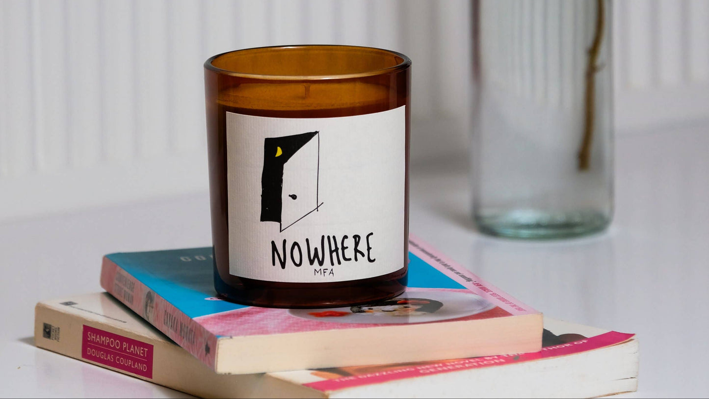 An amber glass candle jar with a white label reading NOWHERE by R.A, styled on top of stacked books with a clear glass bottle in the background against a white paneled wall.