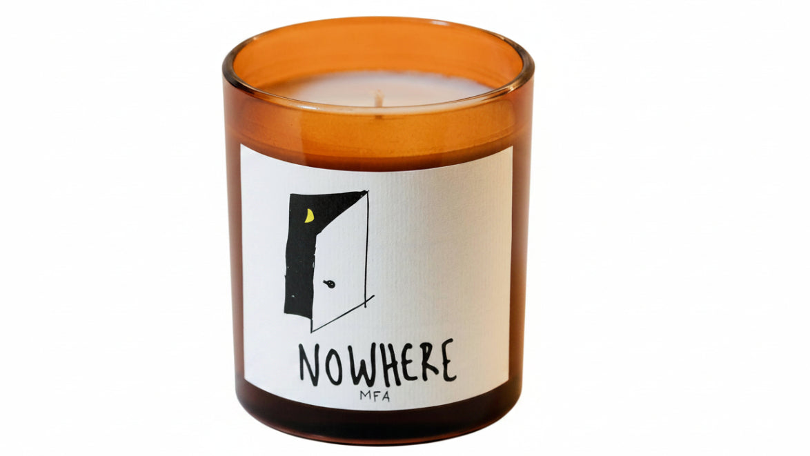 An amber glass jar candle with a white label featuring a minimalist black line drawing of an open door with a small yellow moon and the text 'NOWHERE MFA'.