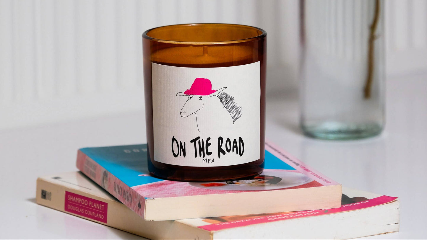 Candle with 'On The Road' label on books next to a clear bottle against a white background