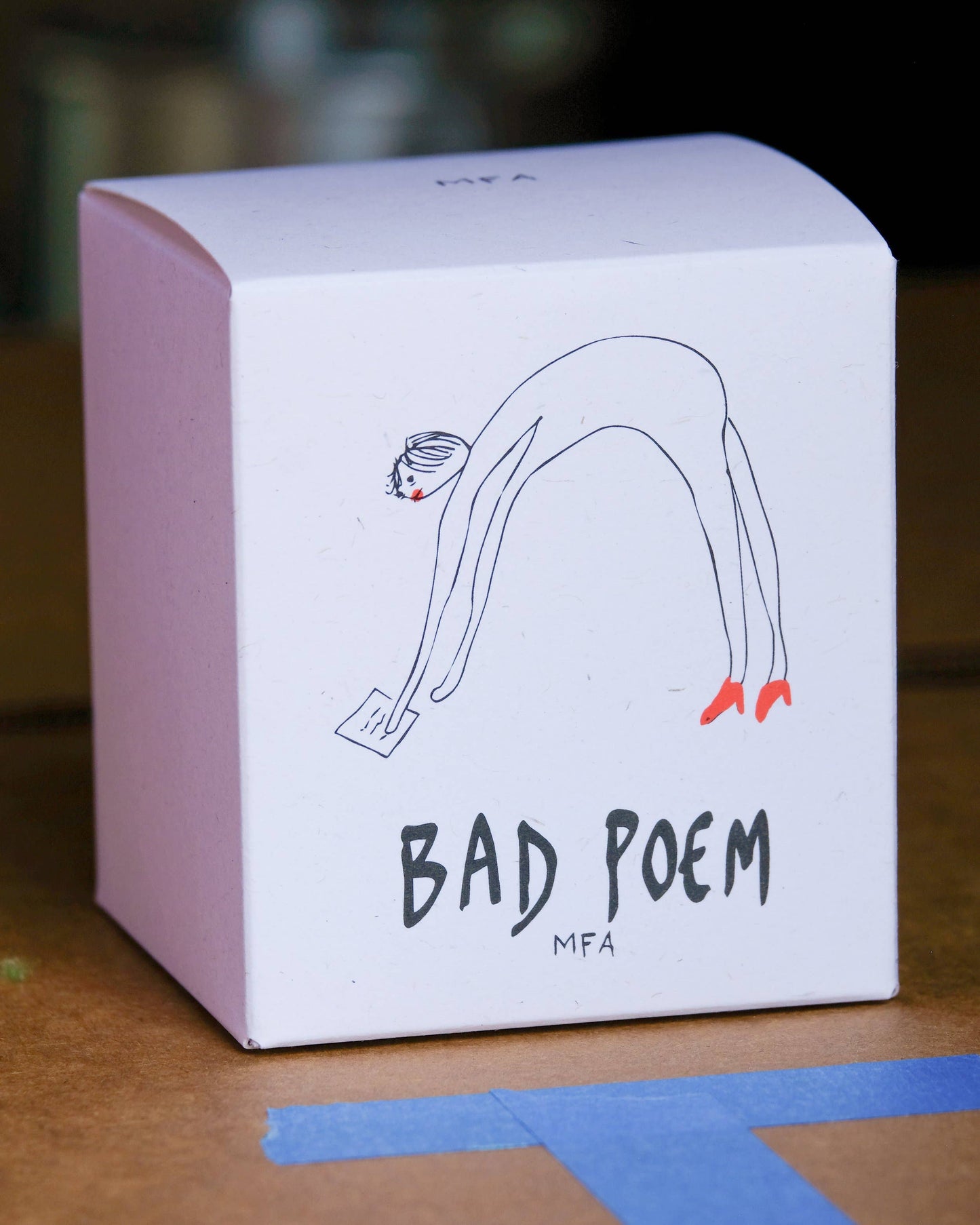 A white square product box with a minimalist line drawing of a person bending over and the text 'BAD POEM' and 'MFA' printed on the front, photographed on a wooden surface.