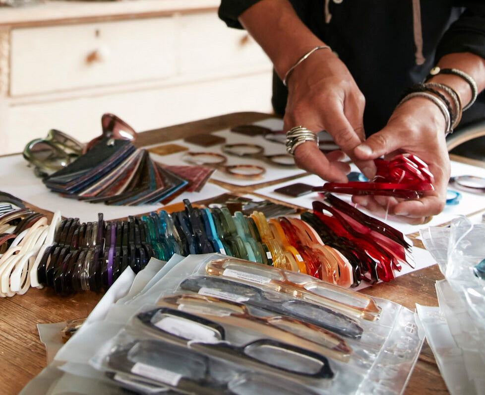 A close-up of a person's hands selecting translucent red acetate frames from a display of various colored eyeglass frames on a wooden table.