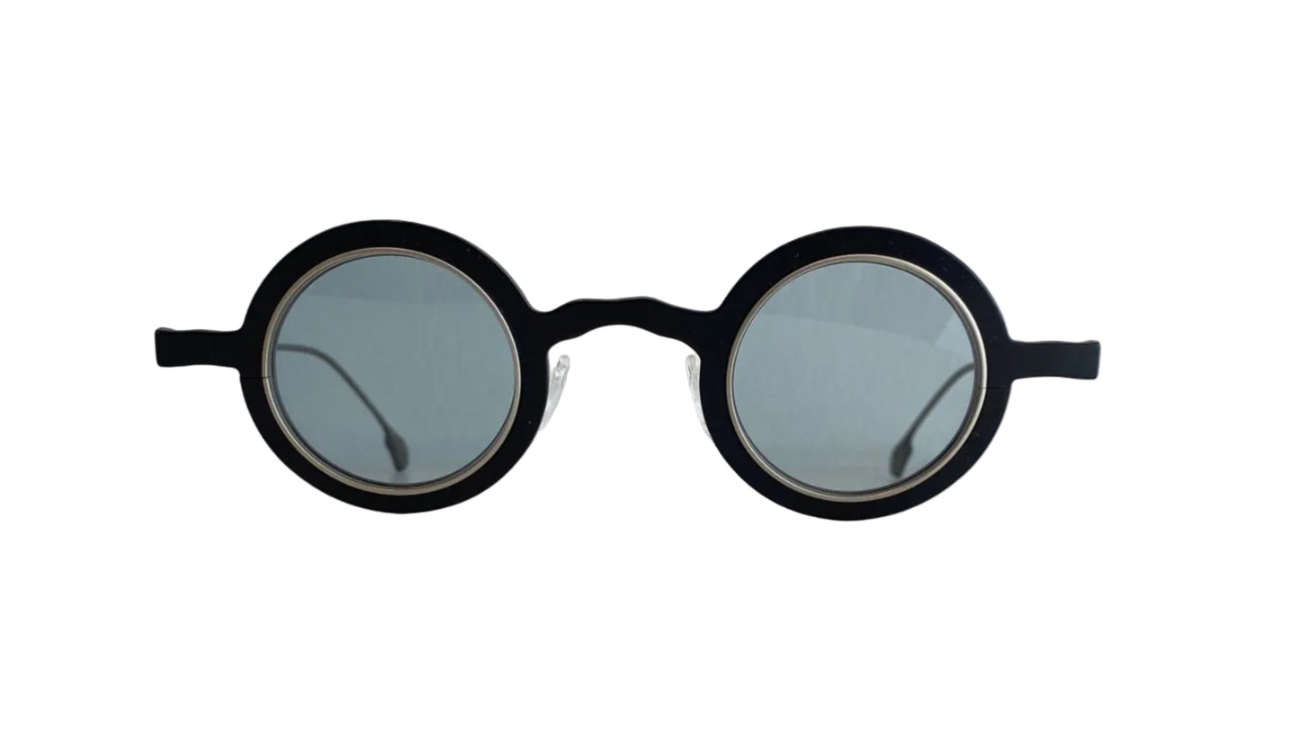 Round black sunglasses with light gray lenses resting on a white surface against a white wall.