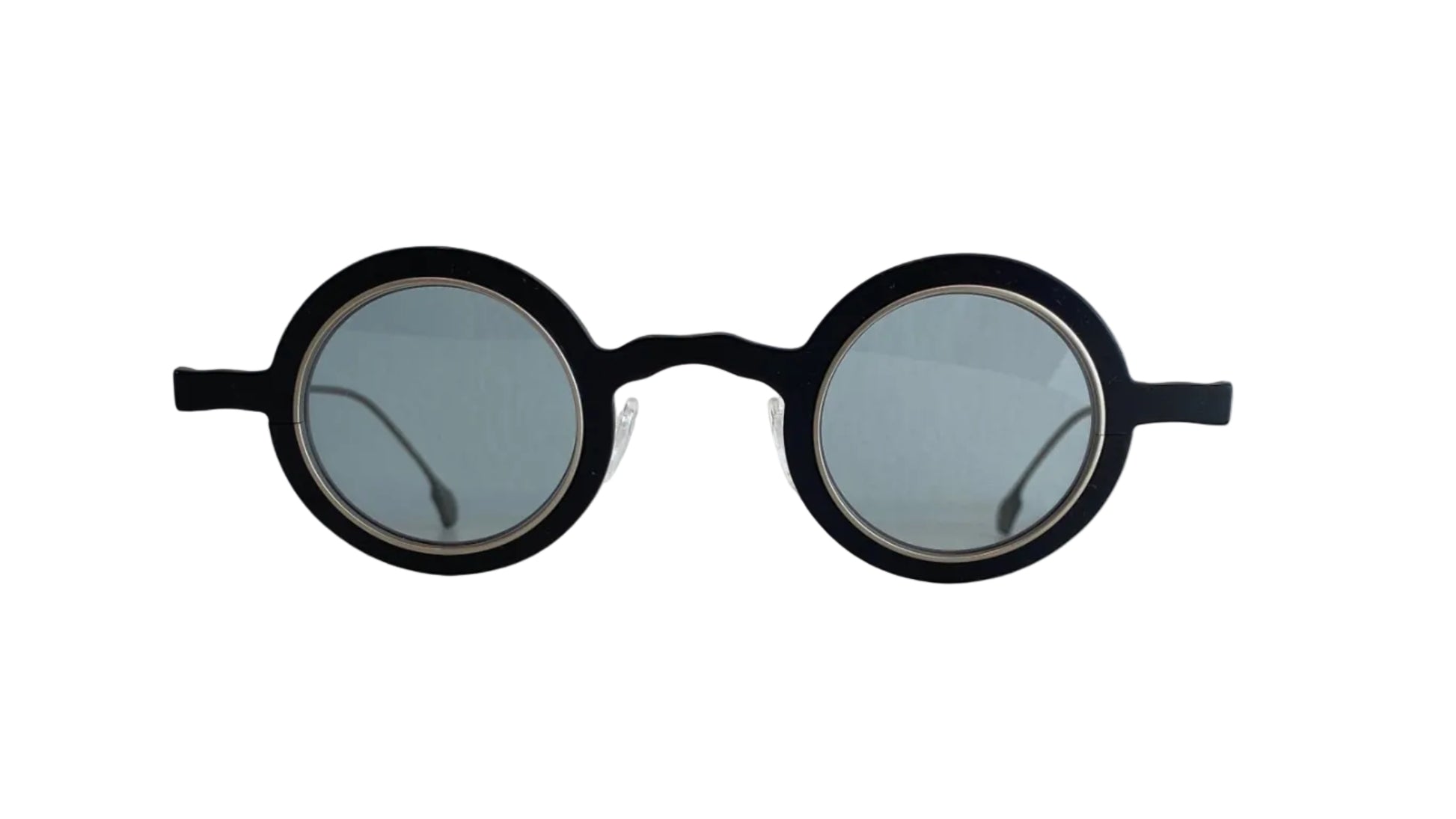 Round black sunglasses with light gray lenses resting on a white surface against a white wall.