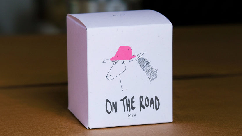 A white cardboard box with a hand-drawn illustration of a white horse wearing a bright pink hat and the text 'ON THE ROAD' and 'MFA' printed on the front, photographed on a wooden surface with a blurred background.