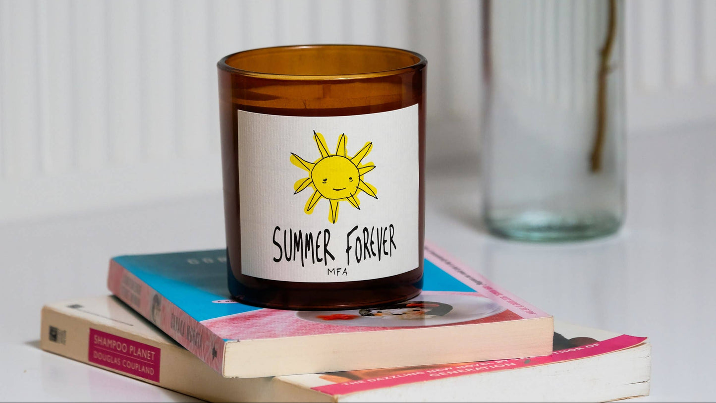 An amber glass candle jar with a white label featuring a yellow sun and the text 'SUMMER, FOREVER' and 'PFAA', styled on top of stacked books with a blurred glass bottle in the background.