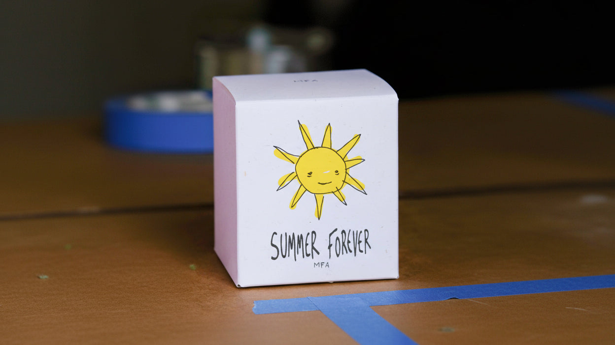 A white square product box with a hand-drawn yellow sun and the text 'SUMMER FOREVER MFA' on the front, photographed on a wooden surface with a blurred indoor background.